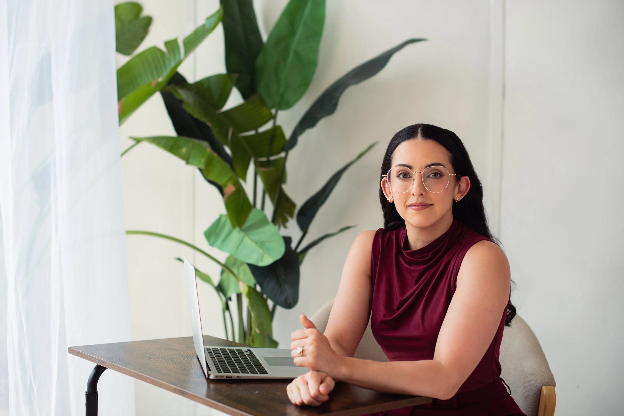 A woman with dark hair and glasses sitting at a wooden desk with a laptop, wearing a sleeveless maroon dress, in a room with large green plants and white walls.