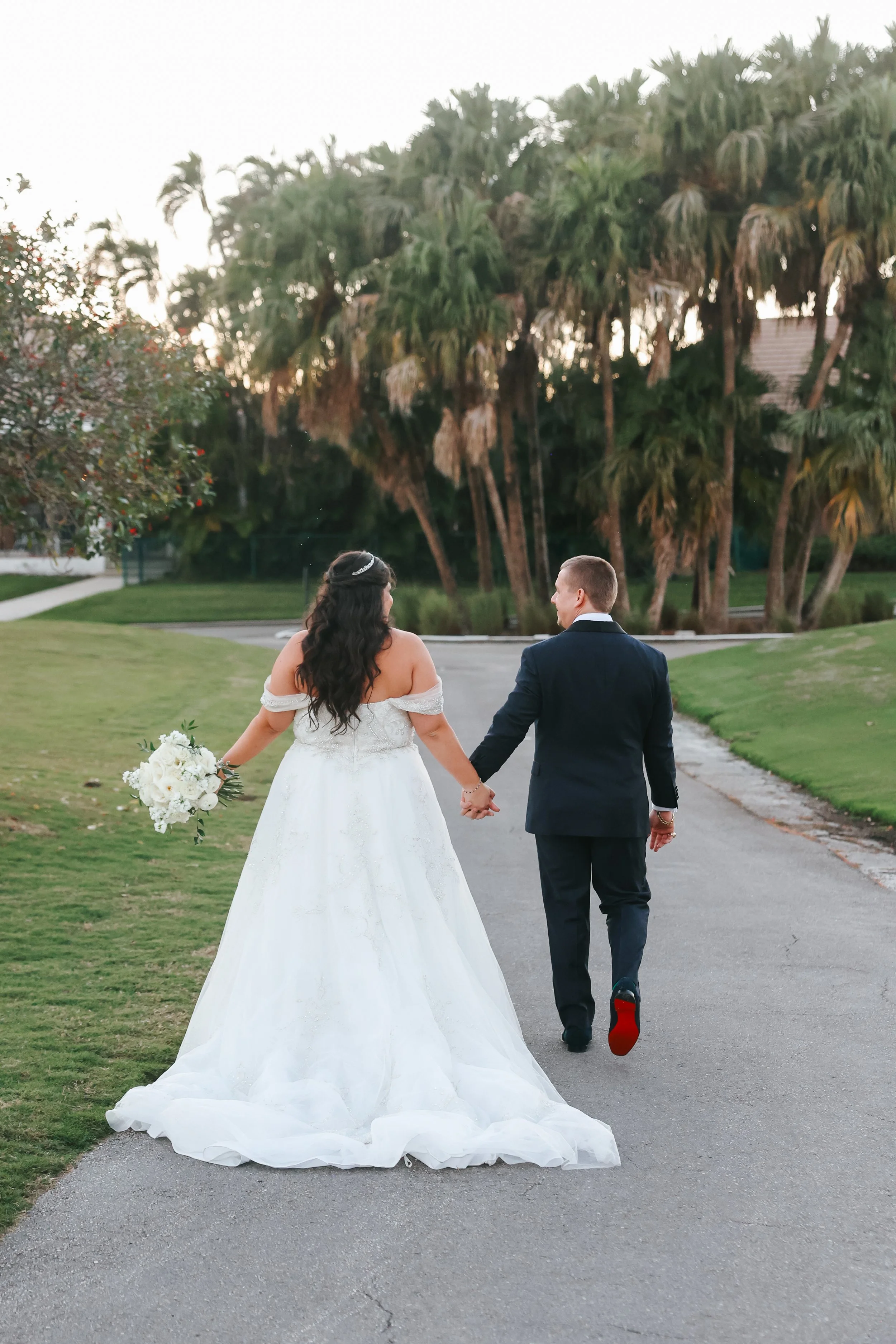 A bride in a white wedding gown holding a bouquet and a groom in a dark suit holding hands as they walk on a paved pathway in a park with green grass, trees, and tall palm trees in the background during sunset.