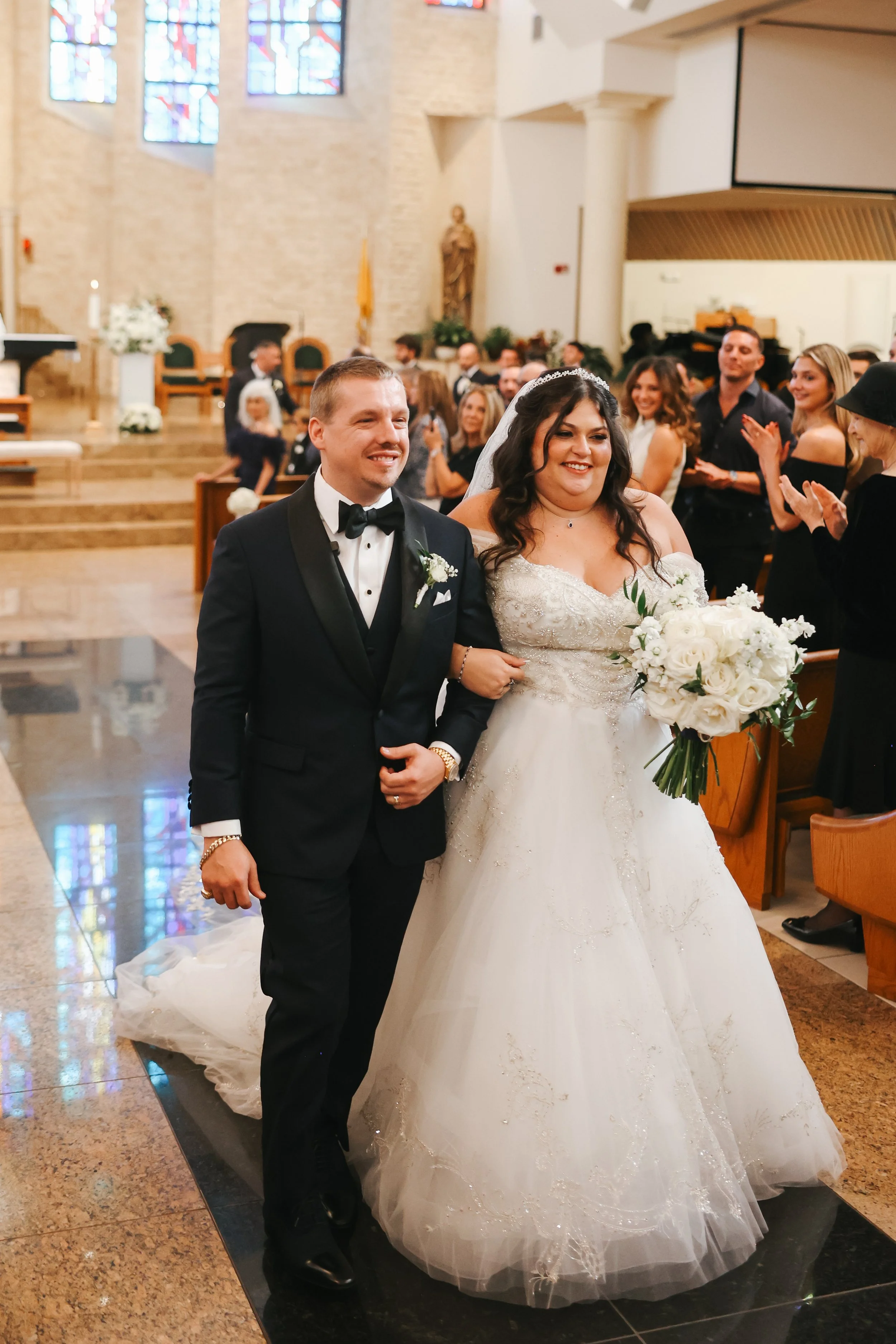 A newly married couple walking down the aisle in a church. The groom is wearing a black tuxedo and the bride is in a white wedding gown holding a bouquet of white flowers, smiling.