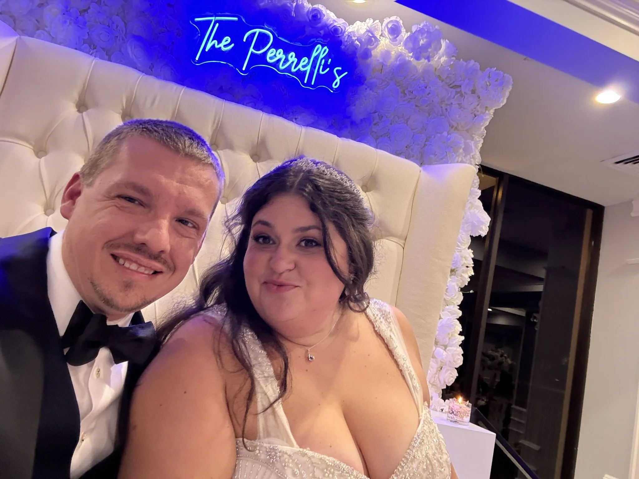 A smiling man in a tuxedo and a woman in a white dress taking a selfie at a wedding reception with a floral backdrop and a neon sign that says 'The Peerelli's'.