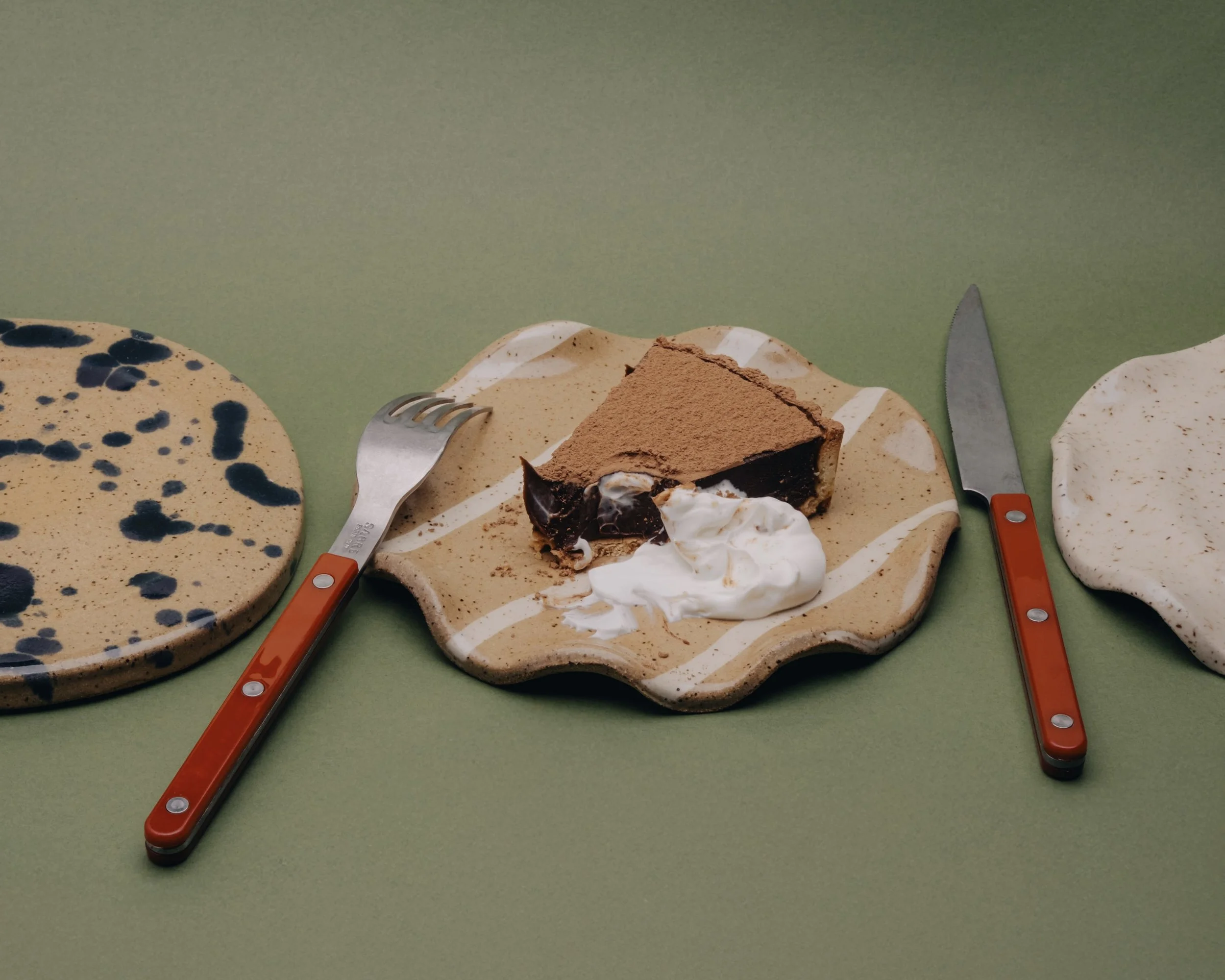 Three decorative plates with desserts on a green surface, with butter knives. The plate on the left has black and beige speckled patterns. The middle plate has a slice of chocolate mousse cake with whipped cream. The plate on the right has white and beige speckled patterns.