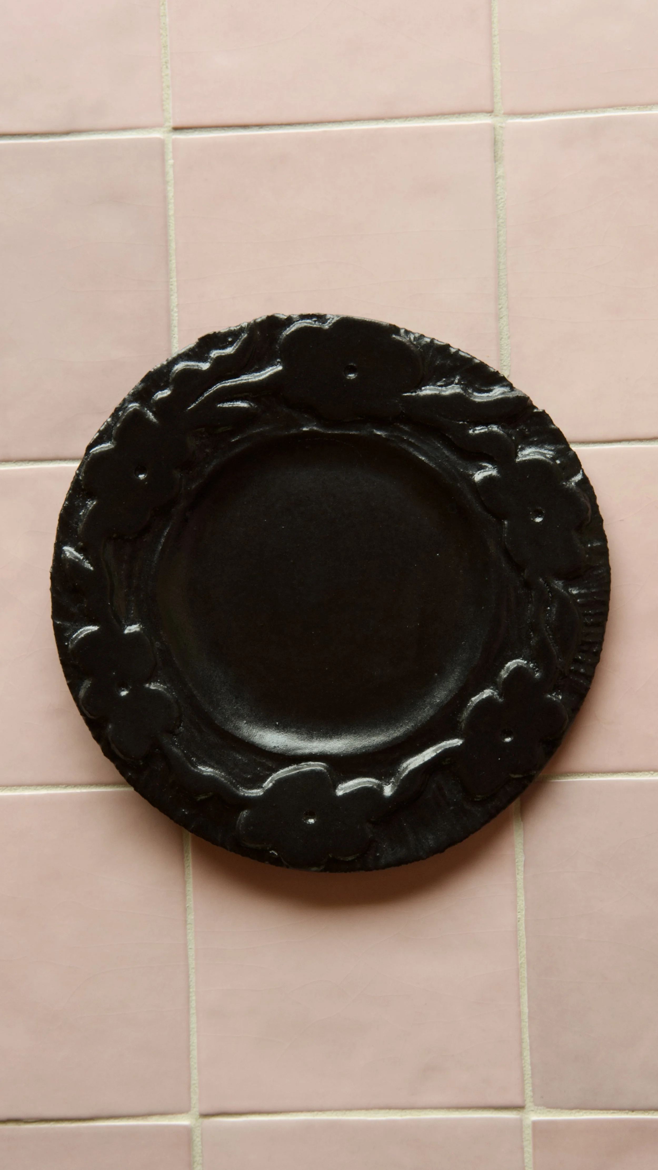 Black decorative plate with a floral border pattern, placed on pink tiled floor.