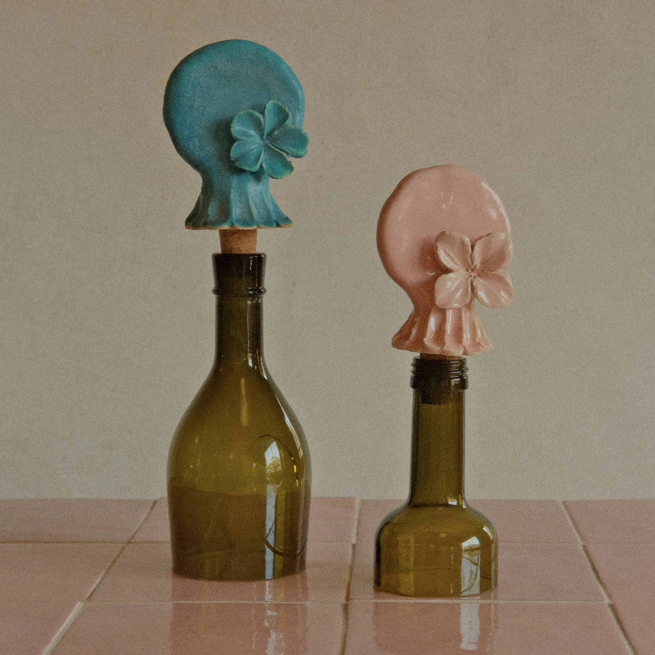 Three glass bottles with decorative soap bars on top, placed on a tiled floor against a plain wall. Each soap bar is shaped like a flower or a face with a floral decoration, in pastel colors.