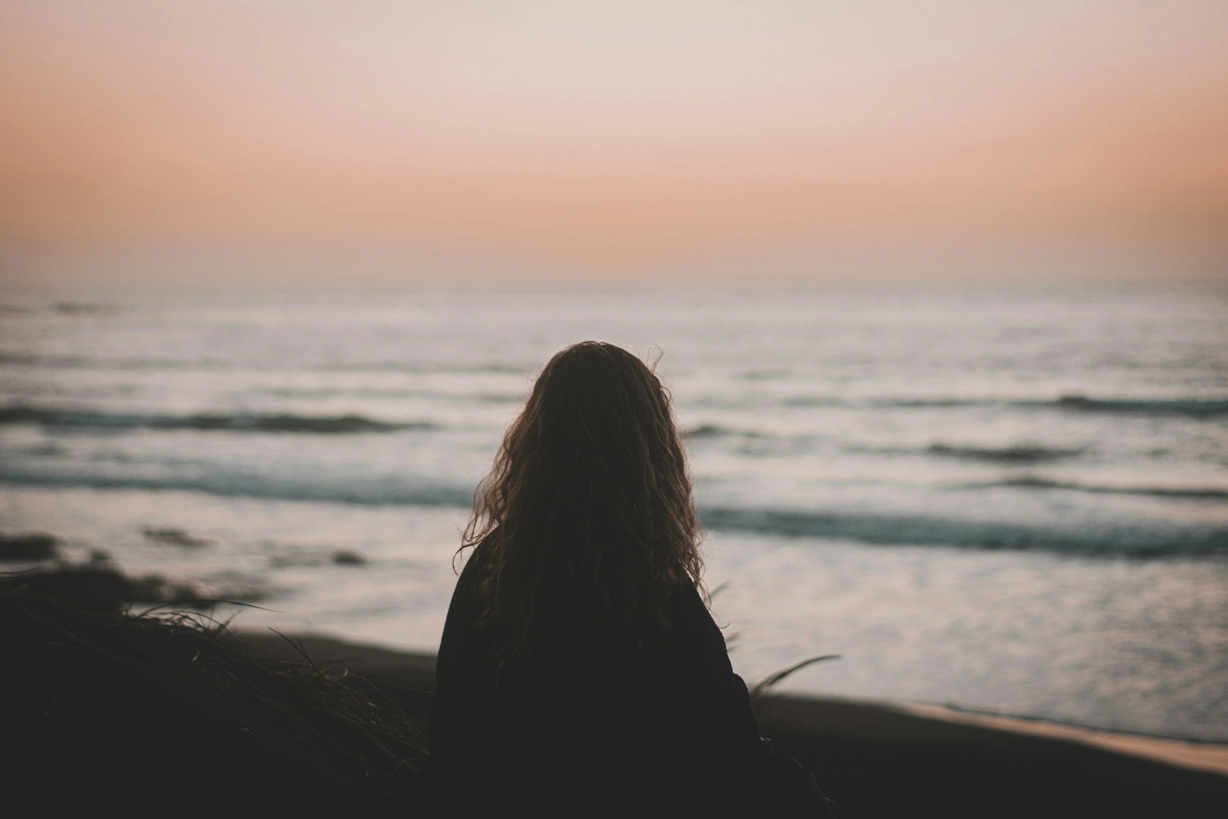 A person with long dark hair sitting on the beach, facing the ocean during sunset or sunrise.