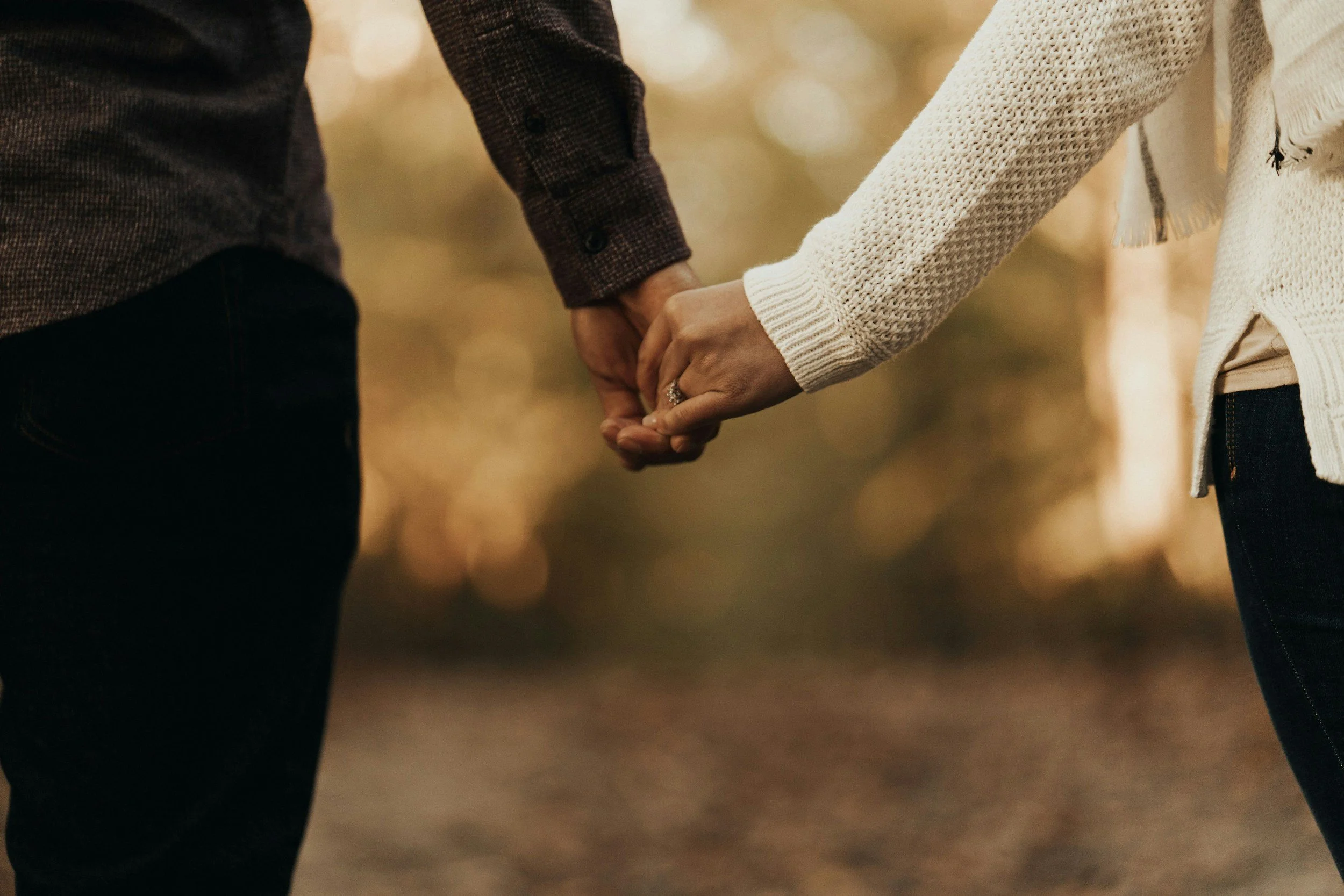 Two people holding hands outdoors, one wearing a white knitted sweater and the other a dark plaid shirt, with blurred autumn background.