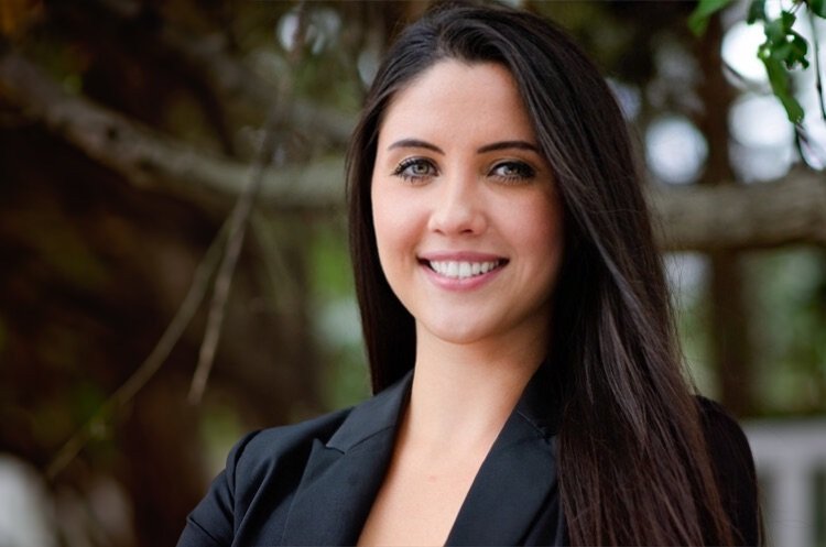 A young woman with long dark hair smiling outdoors, wearing a black blazer with a blurred natural background.