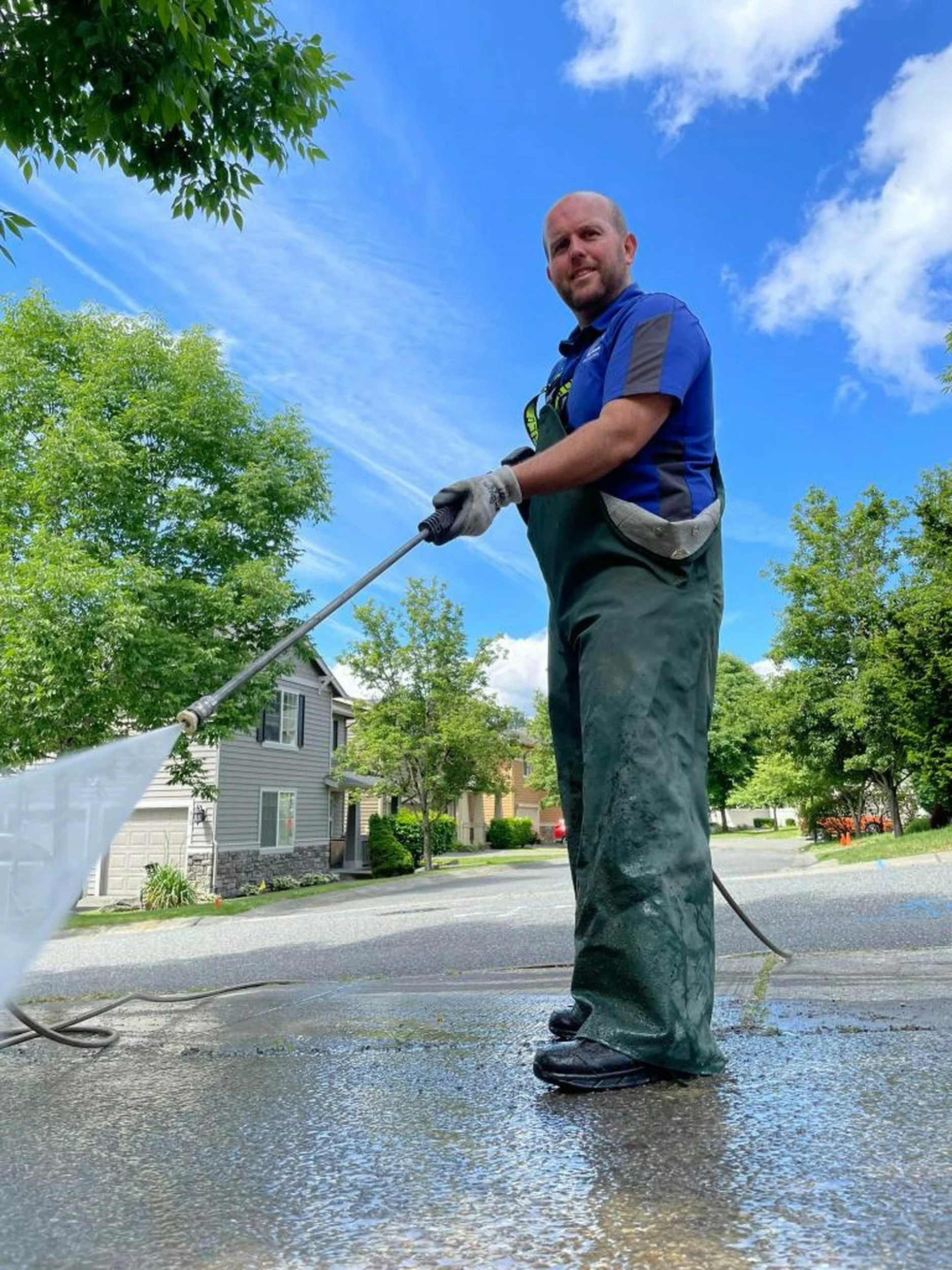 A man wearing a blue shirt, gray gloves, and black rubber boots is pressure washing a roof on a residential street. The scene is outdoors with a blue sky, white clouds, and leafy green trees in the background.