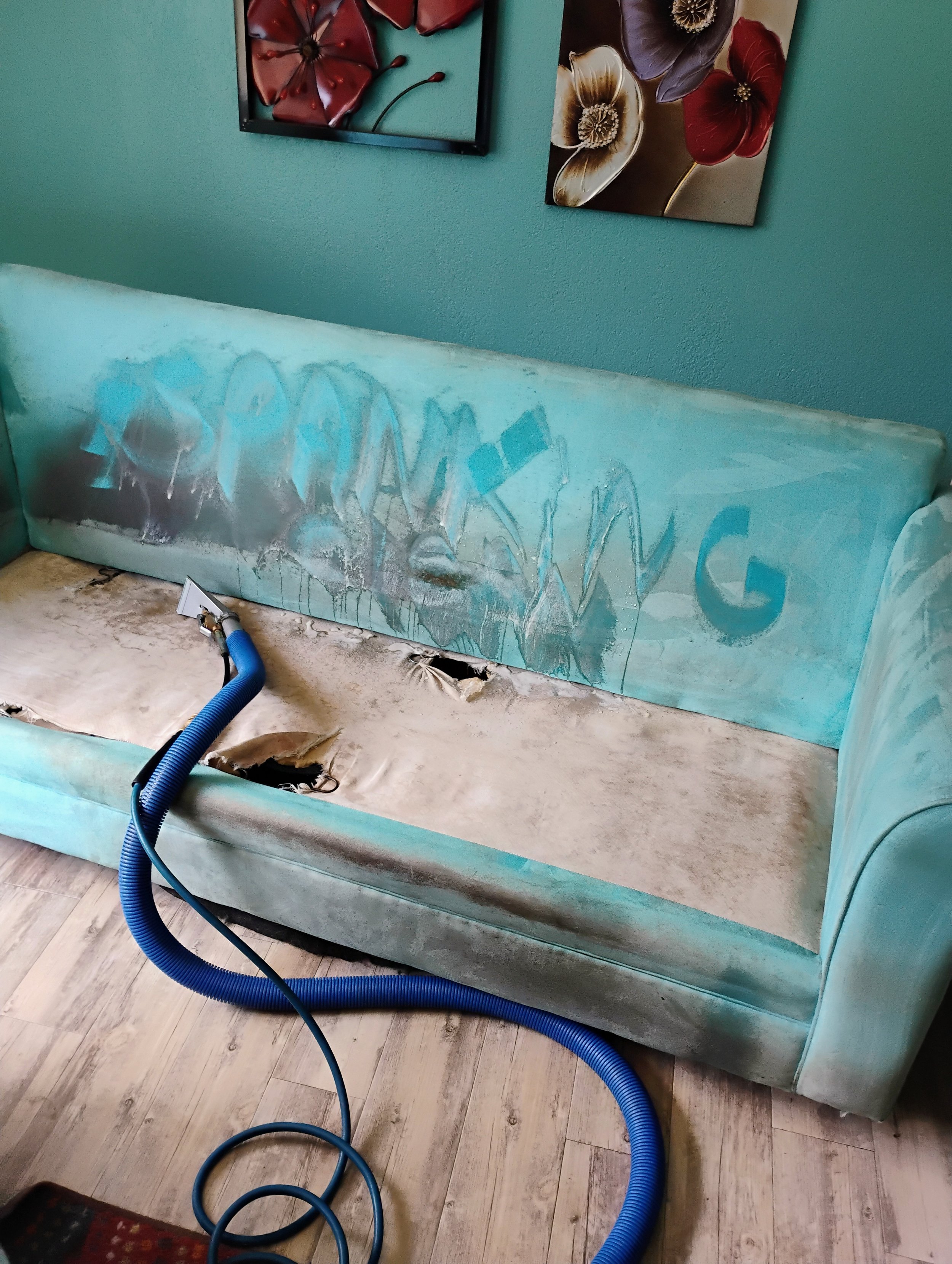The image shows a partially stripped teal-colored couch with discoloration and graffiti on the fabric. There is a large blue vacuum hose connected to equipment near the couch, implying cleaning or restoration work. Behind the couch, there are two pieces of floral artwork on the wall, and the floor is made of light-colored wood planks.