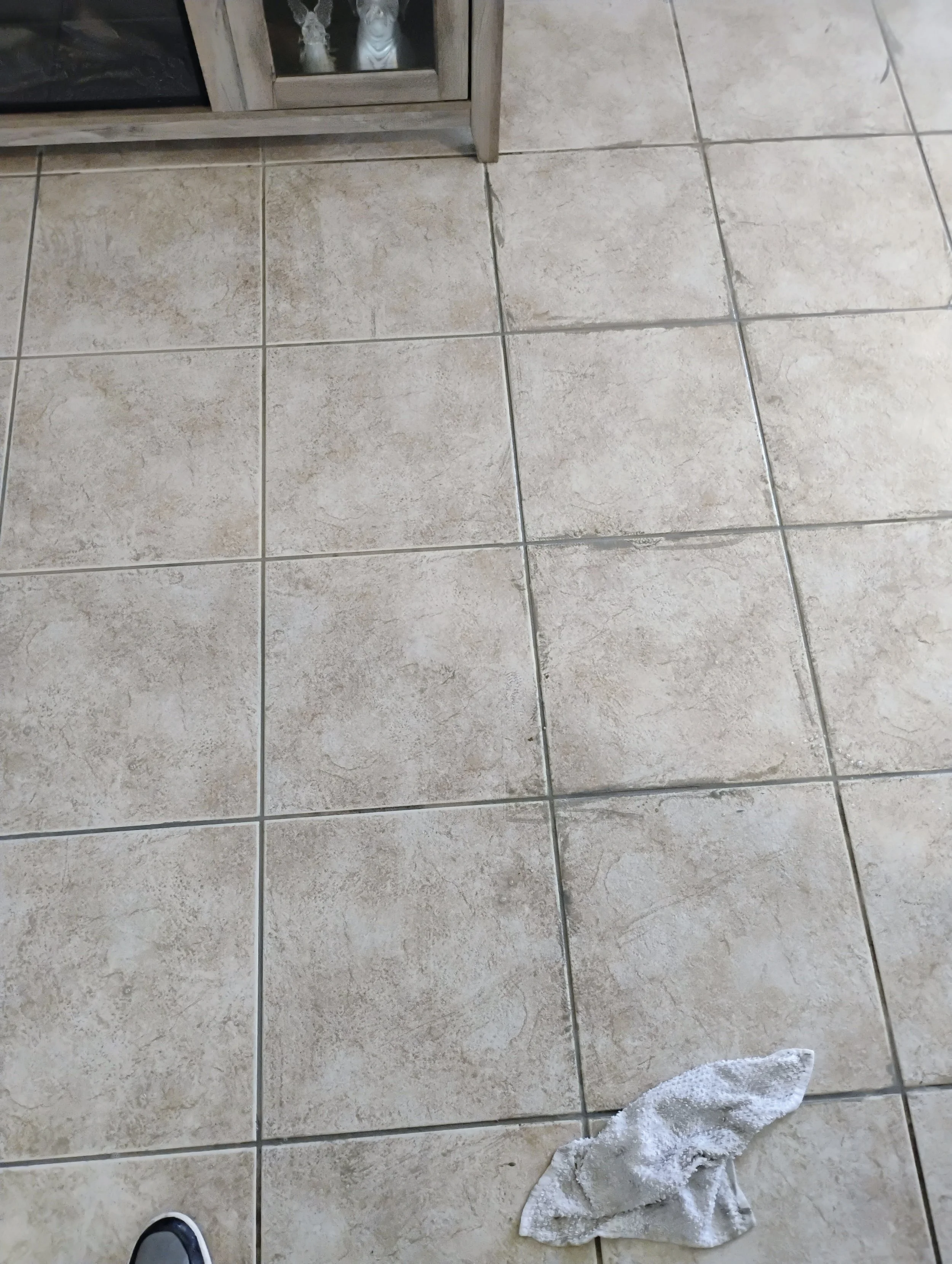 A tiled floor with beige and brown tiles, a white cloth towel, and a person's foot wearing a blue and white shoe.