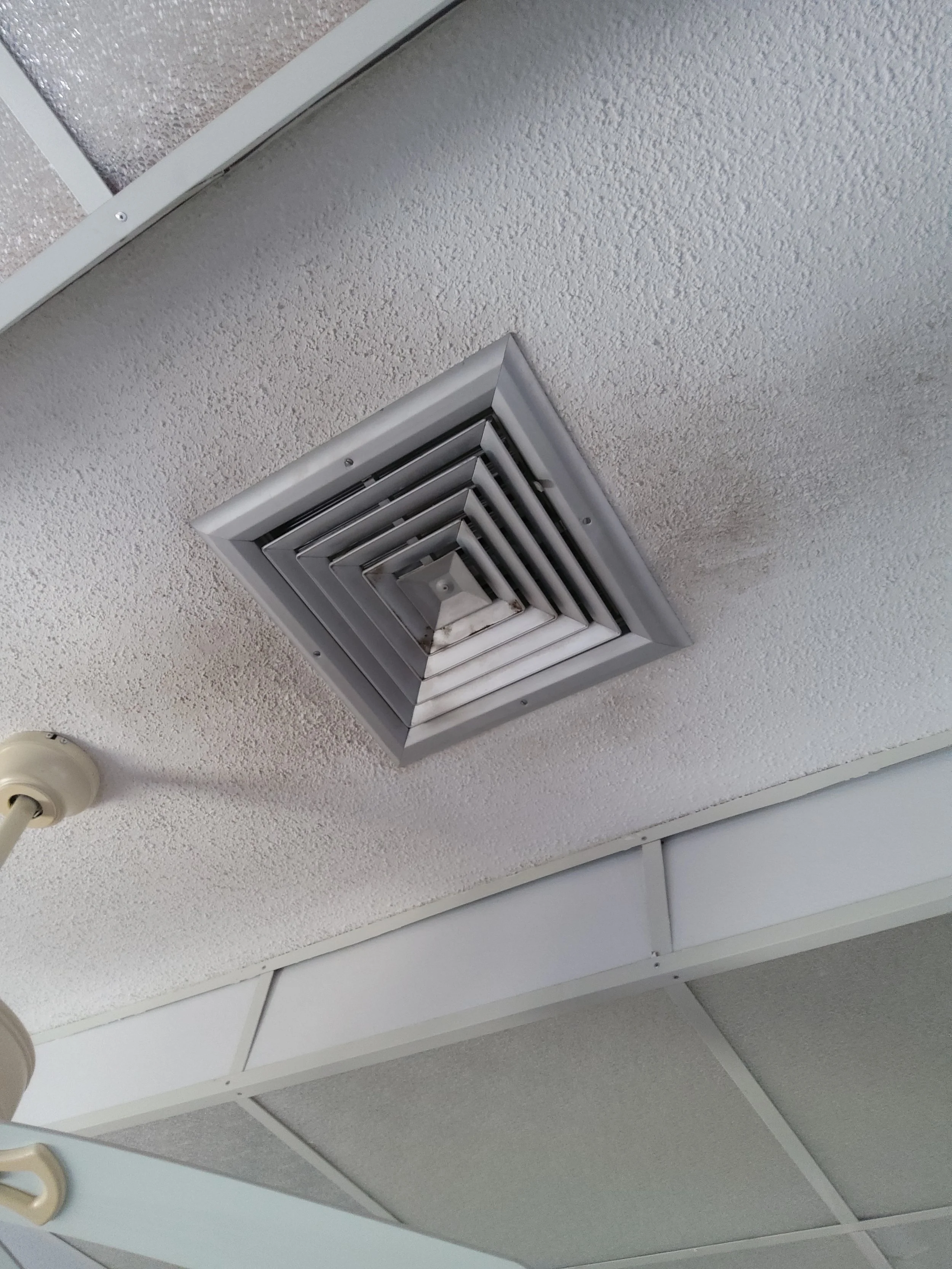 Close-up view of a ceiling with a square air vent and a round smoke detector or sprinkler near the edge.