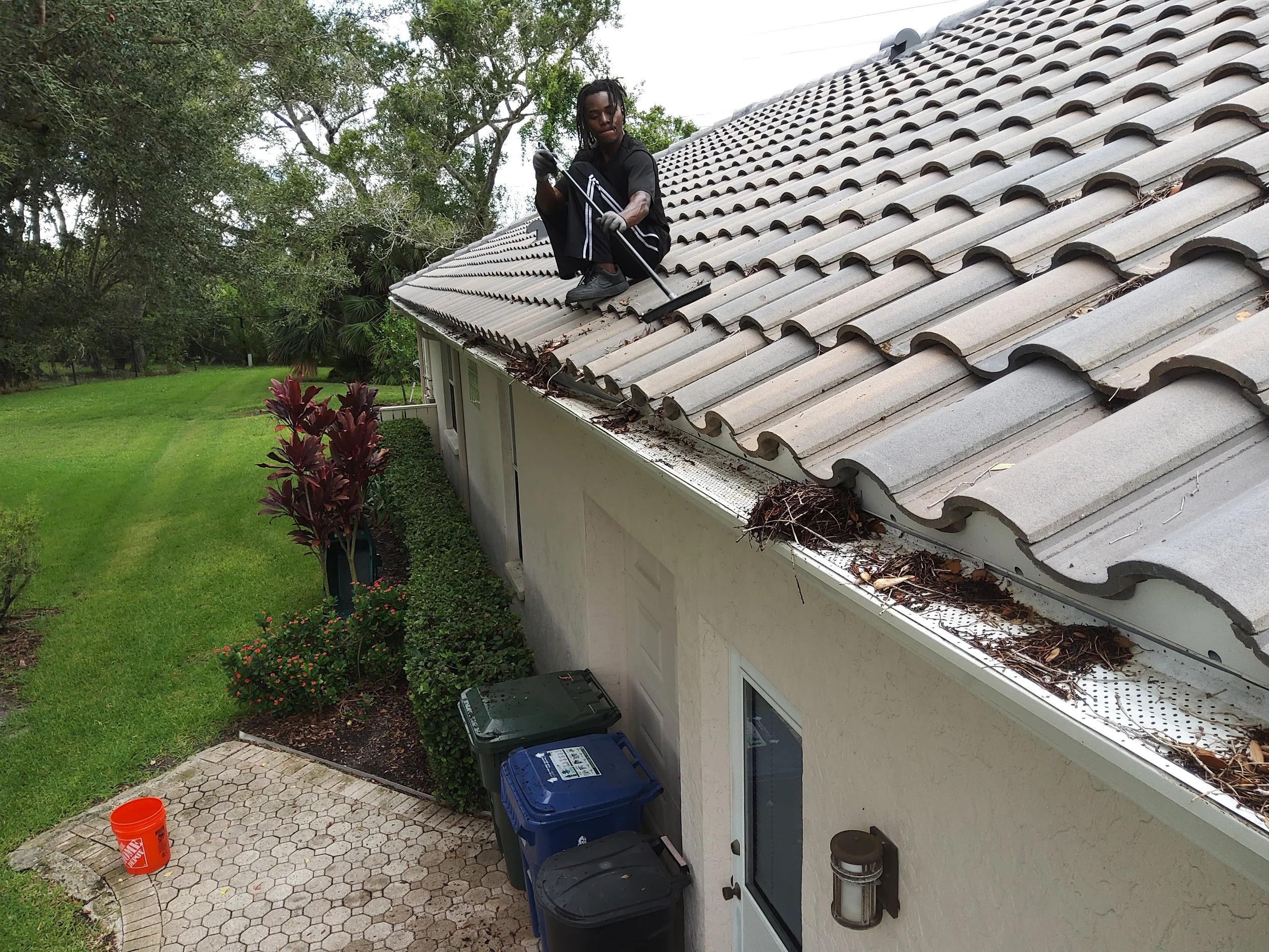 A man on a roof, wearing black, working with a tool, with trees and a grassy yard in the background, and trash bins and a patio below.