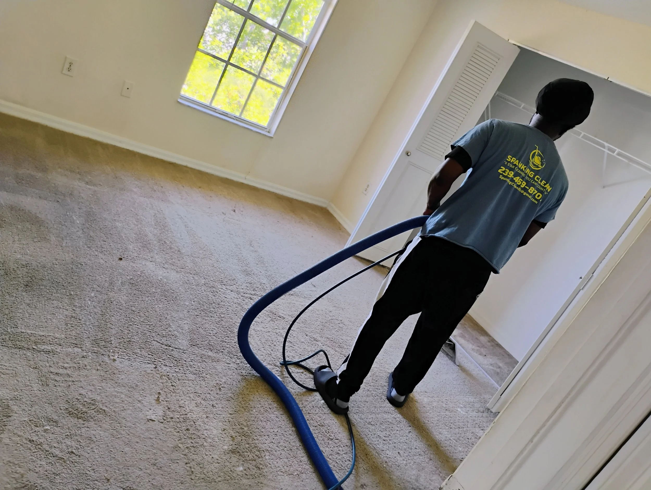 A person vacuuming a beige carpet in a room with a large window showing green trees outside. The person is wearing a blue shirt with yellow text and black pants.