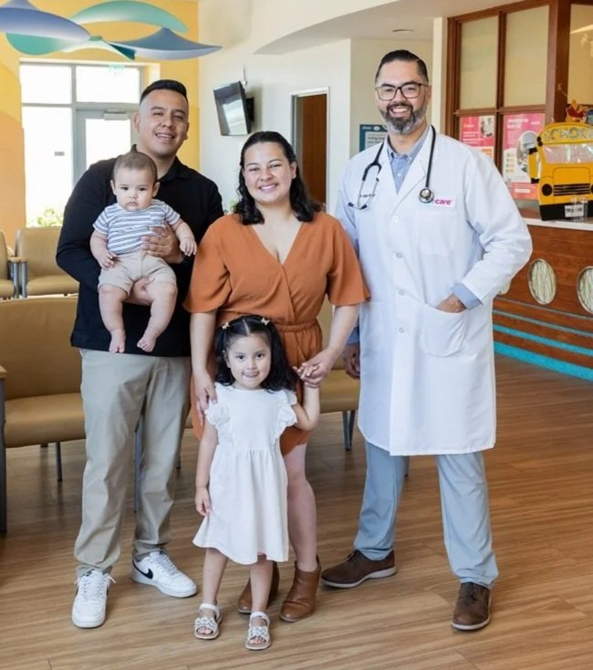 A healthcare professional and a family of four pose together in a medical facility. The family includes a man holding a baby and a woman with a young girl, all smiling happily. The background features colorful decorations and seating.