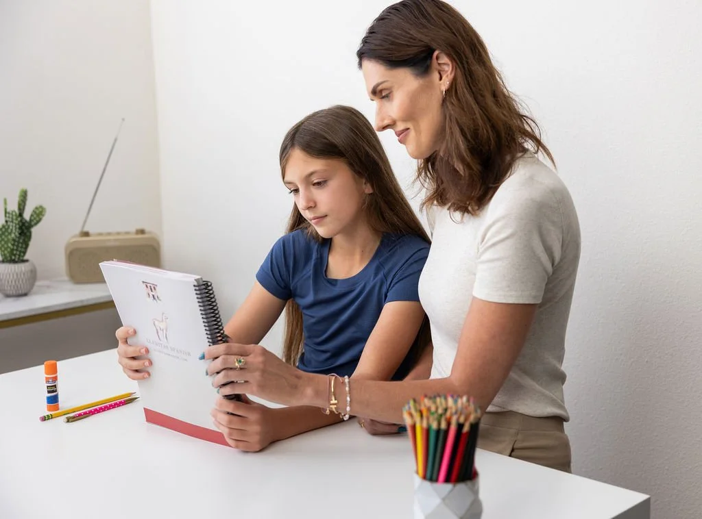 A woman and a girl sitting at a white table, looking at a notebook. The table has colored pencils, a glue stick, and a container of pencils. There is a plant and a vintage radio in the background.