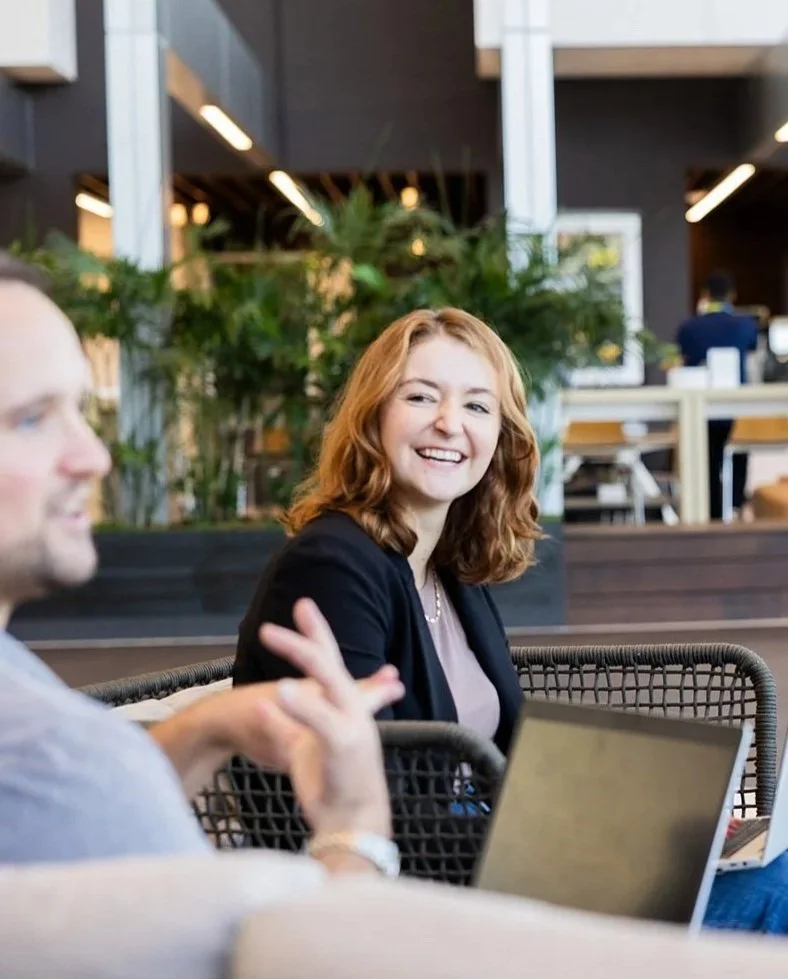 A woman with red hair smiling and looking at a man who is talking, in a modern indoor space with greenery and people working in the background.
