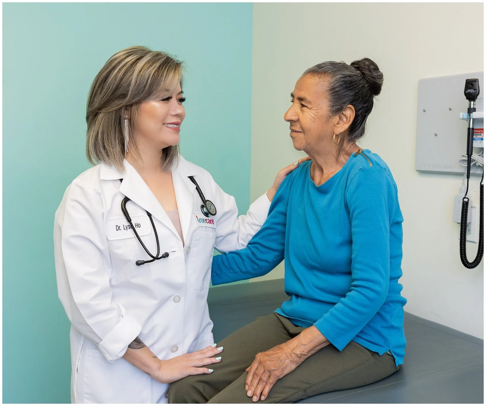 A healthcare professional, a female doctor, with a stethoscope around her neck, is smiling and talking with an elderly woman who is sitting on an examination table. The woman is wearing a blue top and has her hair tied back. They are in a medical examination room.