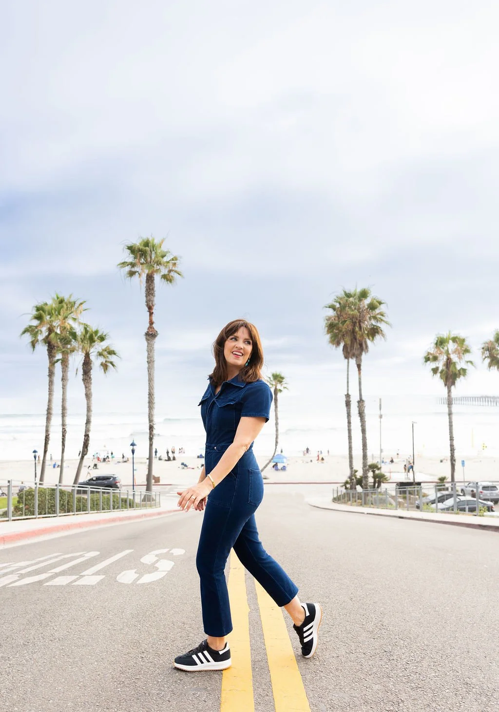 Woman in blue denim jumpsuit and black sneakers walking on street near beach with palm trees and ocean in background.