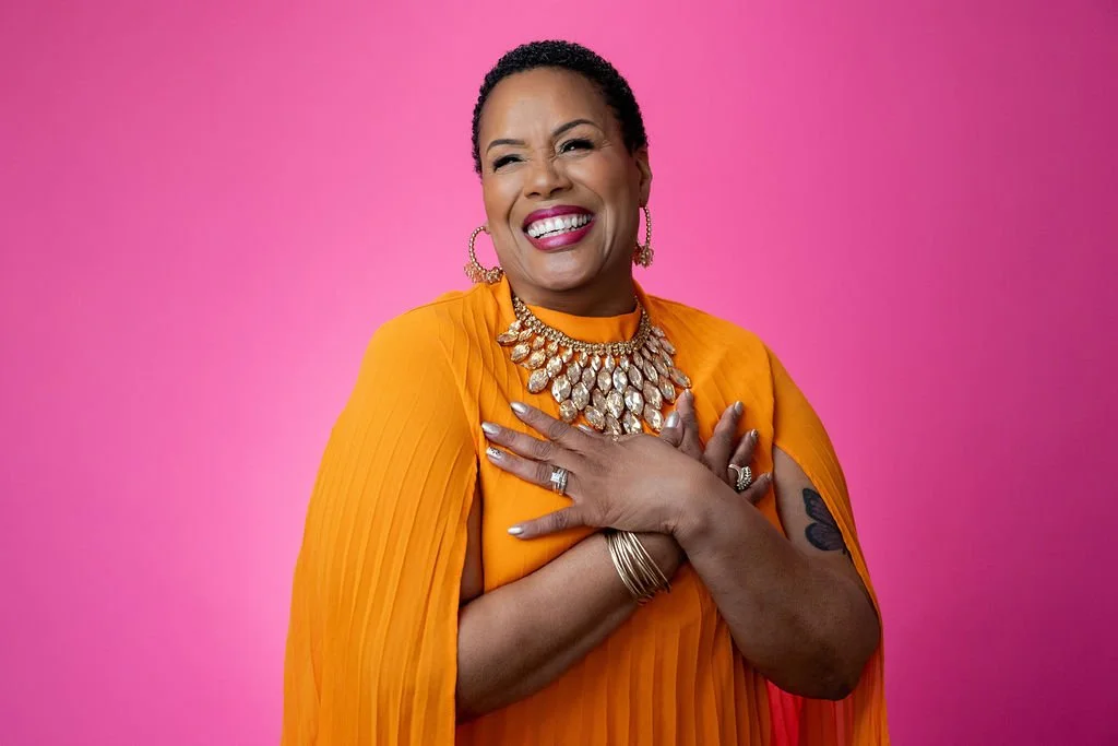 A woman with short curly hair, wearing a bright orange dress and jewelry, smiling with one eye closed against a pink background.