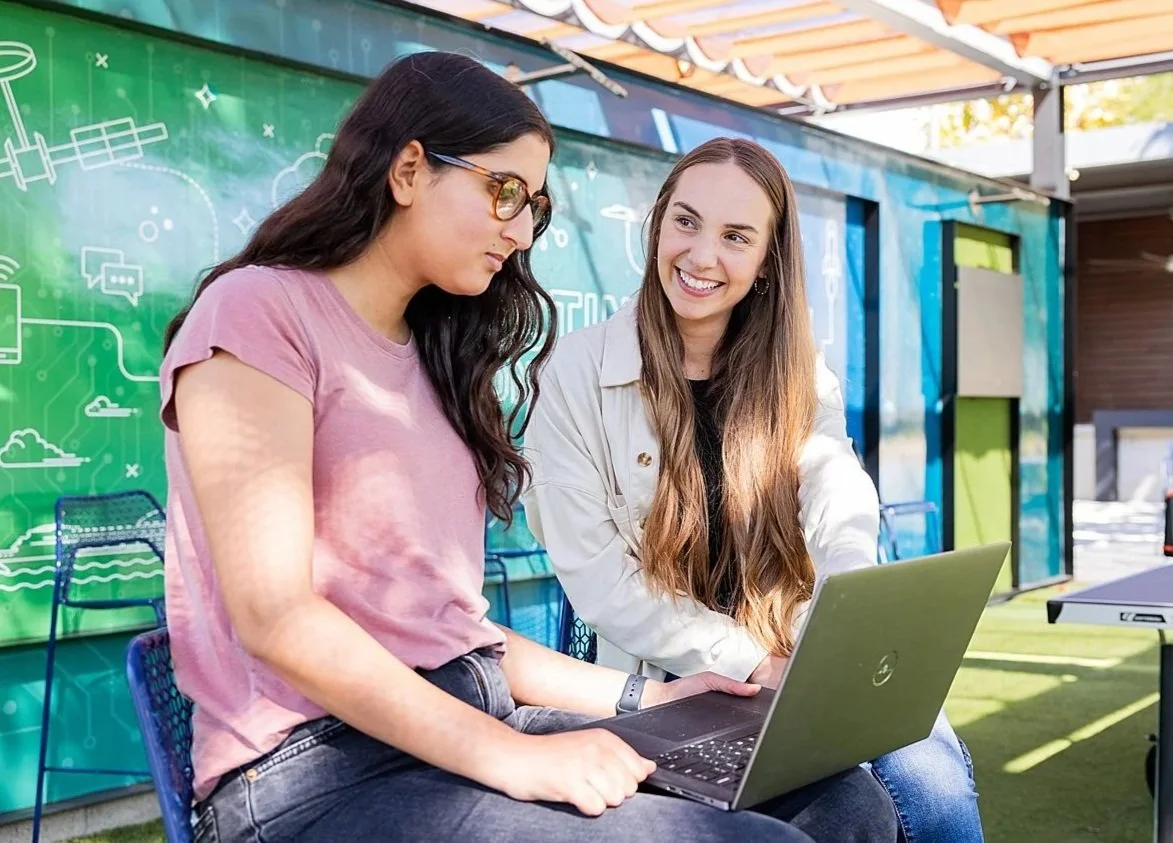 Two women sitting outdoors at a table with a laptop, engaging in a conversation, with a colorful mural in the background.