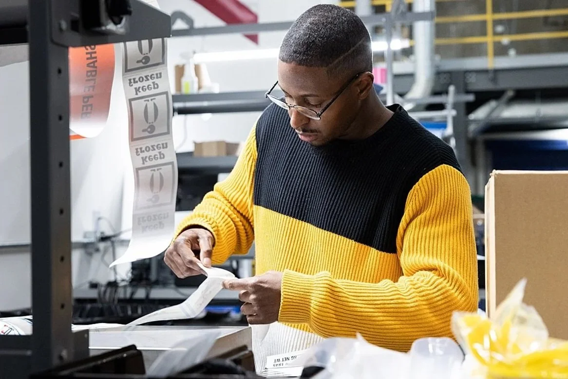 A man with glasses and a buzz cut hairstyle working in a warehouse or packaging facility, handling labels or packaging materials.
