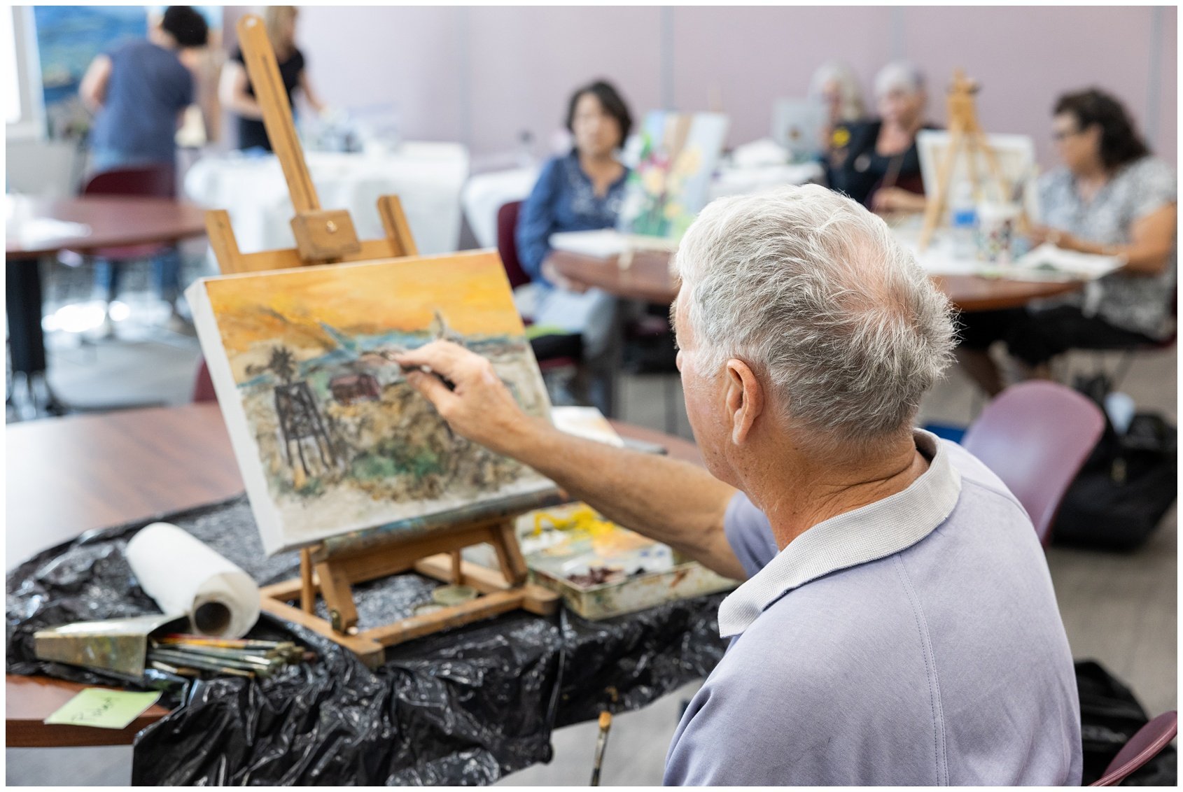An elderly man with gray hair painting a landscape on a small canvas set on an easel at an art class. Several women sit at tables in the background, some using laptops or looking at their paintings, while others are standing and socializing.