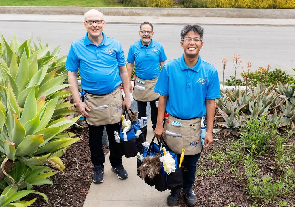 Three smiling workers in blue shirts and beige aprons standing on a sidewalk surrounded by green plants. One is holding a black bag with supplies and a small dog, another also has a bag, and the third has a bag and supplies.
