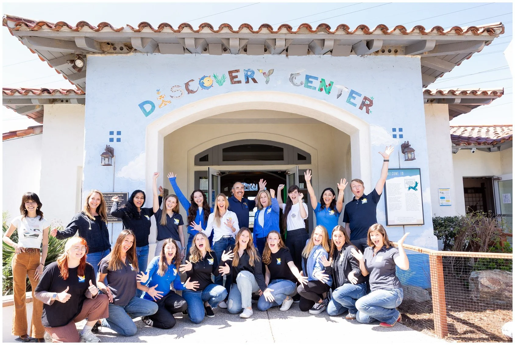 Group of happy people gathered in front of the Discovery Center building, smiling and raising their hands in celebration, with the center's sign and decorative elements above the entrance.