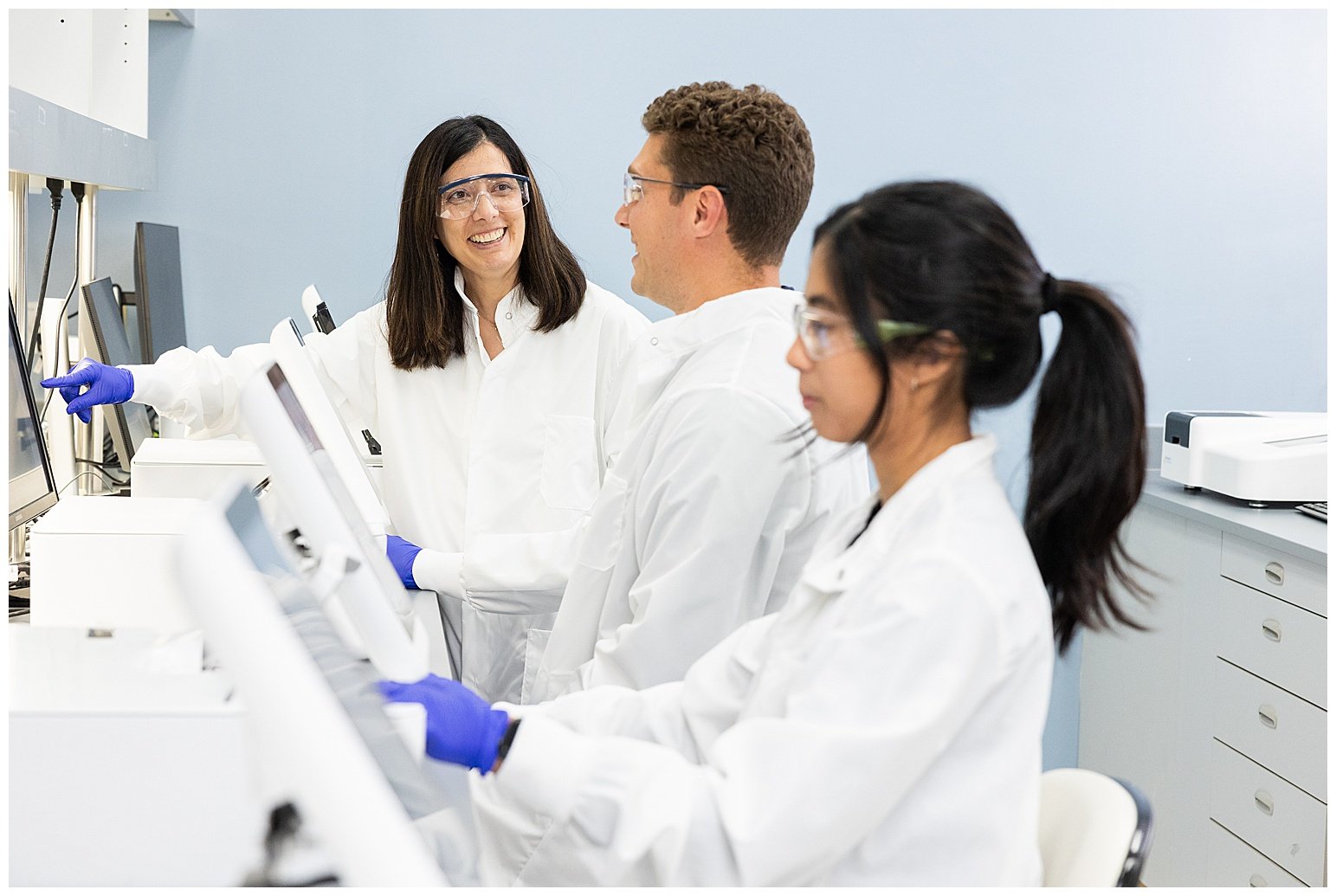 Three scientists in lab coats and safety goggles working in a laboratory with laboratory equipment and computer monitors, engaging in a discussion or demonstration.