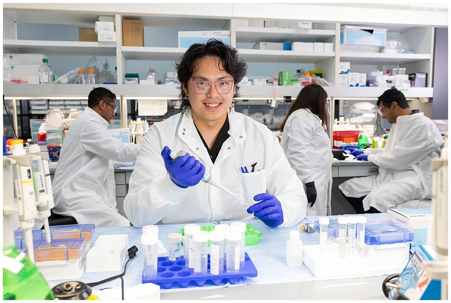 A group of scientists working in a laboratory, with one scientist in the foreground holding a pipette and smiling at the camera, surrounded by lab equipment and supplies.