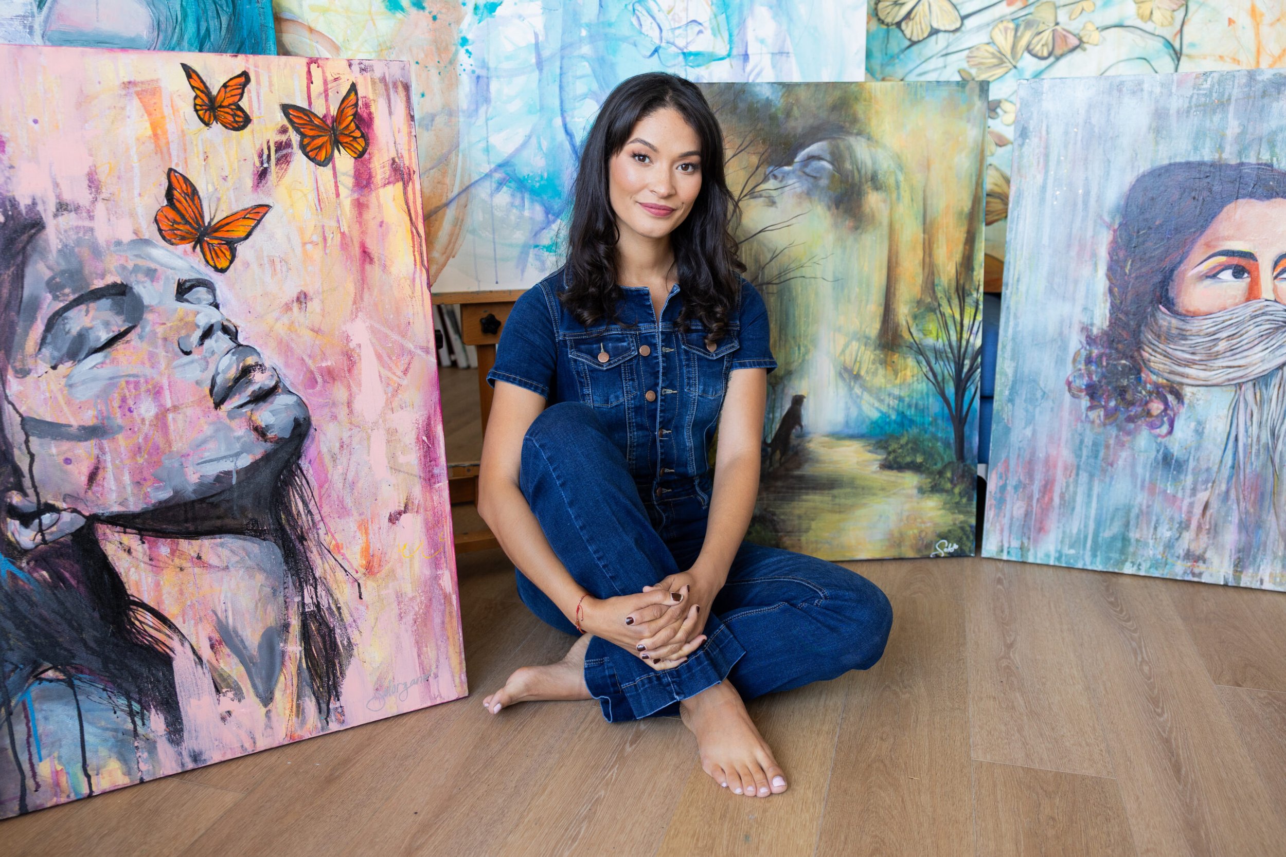 A woman with long dark hair, wearing a denim jacket and jeans, sitting on the floor with her arms crossed in front of her, surrounded by colorful abstract paintings, including butterfly artwork and a portrait of a woman with a face covering.