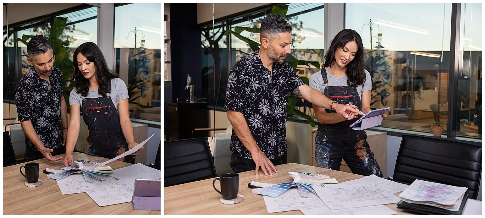 Two people, a man and a woman, are standing at a table in an office, reviewing design plans and color swatches. The man is pointing at a laptop, and the woman is holding a color palette. There are papers, drawings, and a mug on the table, and large w