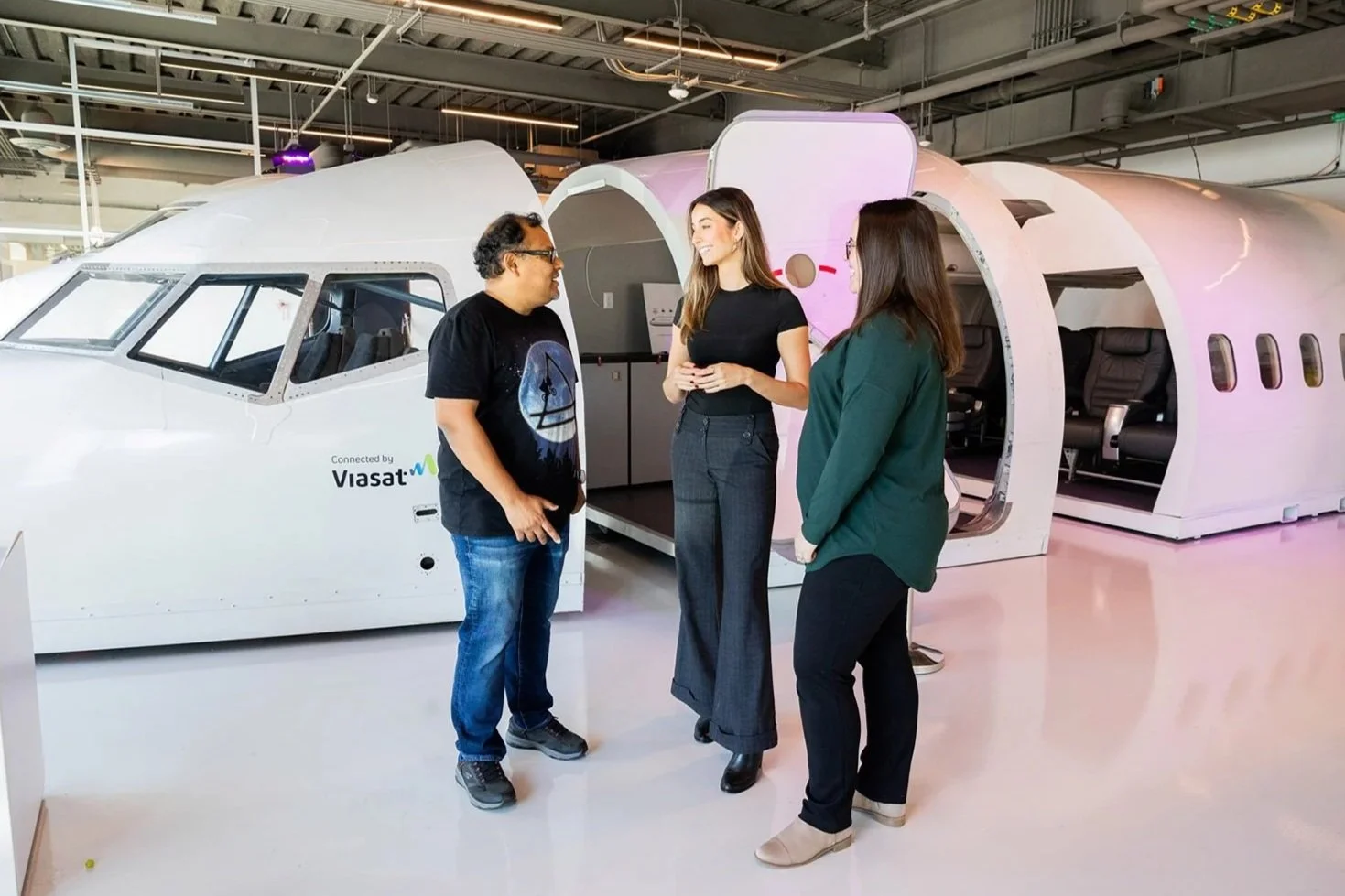 Three people standing and talking inside an aircraft manufacturing or design facility with a futuristic aircraft in the background.