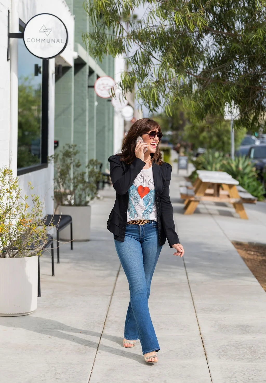 A woman with dark hair, sunglasses, wearing a black jacket, graphic T-shirt, and blue jeans, walking on a sidewalk while talking on her cellphone and smiling.