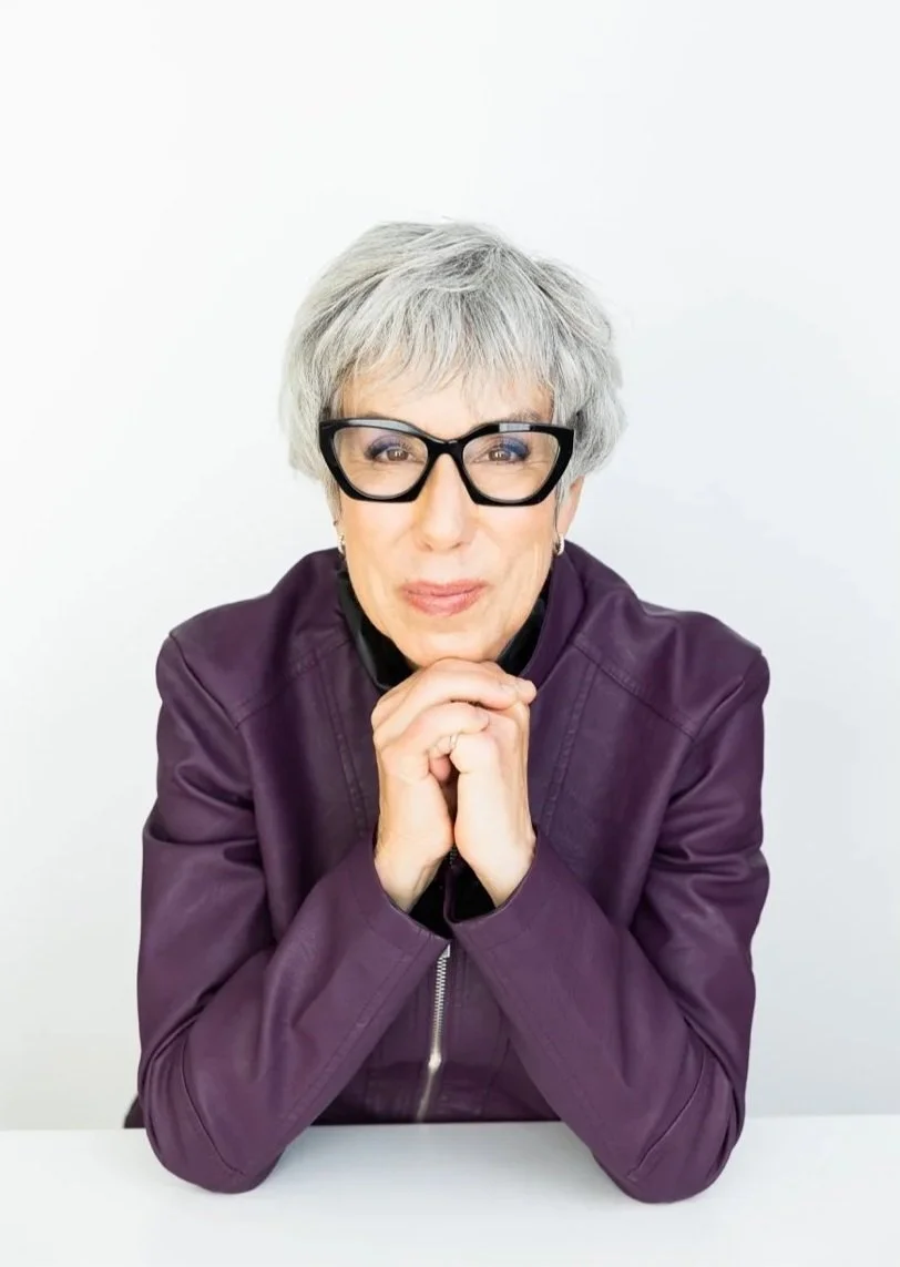 An older woman with short gray hair, large black glasses, and wearing a purple jacket, sitting at a white table with her hands clasped under her chin, looking at the camera.