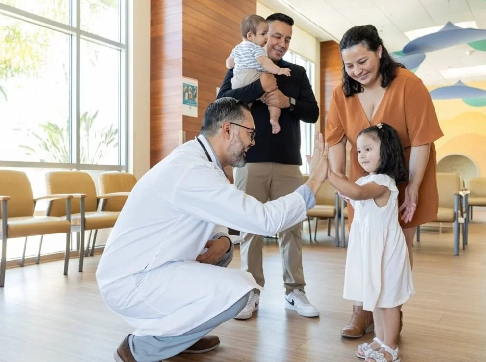 A doctor kneeling and giving a high five to a young girl, her mother assisting, while a family in the background waits in a bright, modern medical office.