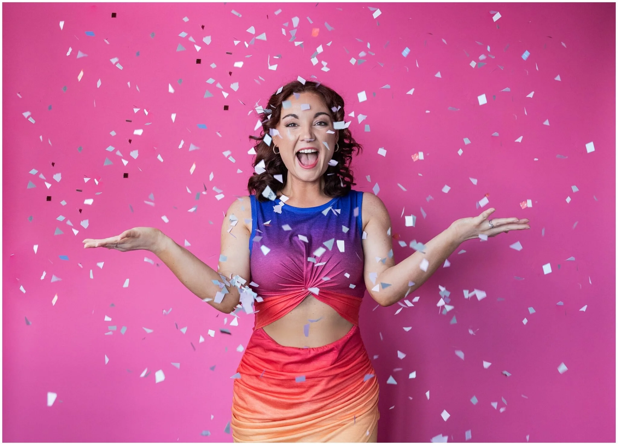A woman celebrating with confetti against a pink background, wearing a colorful tank top and a skirt.