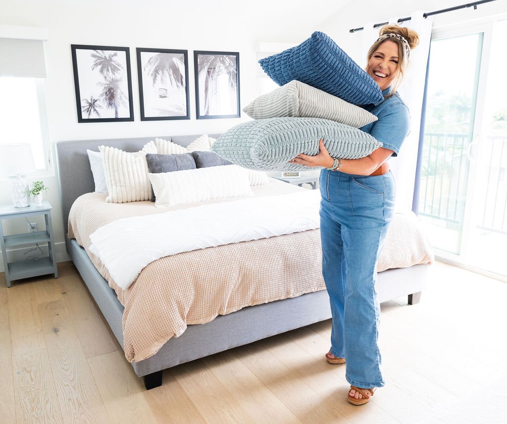 Smiling woman holding four neatly stacked pillows in a bright bedroom with artwork, a bed with pillows, and large windows.