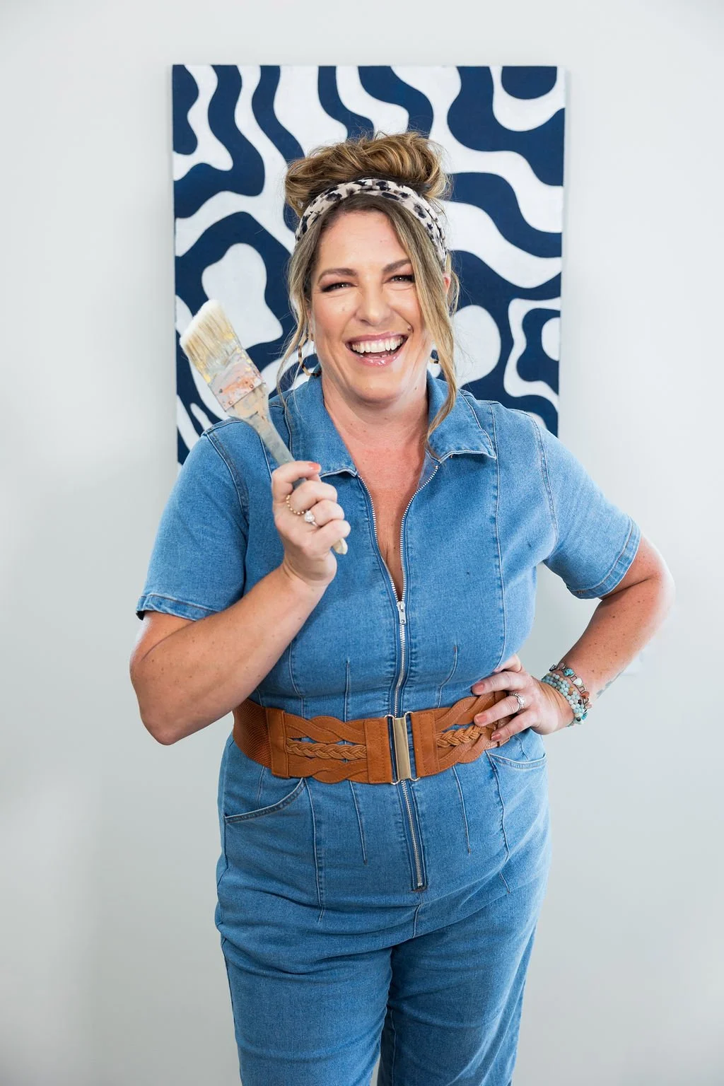 A cheerful woman in a denim jumpsuit and a beige headband, holding a paintbrush, standing in front of a black and white patterned artwork.