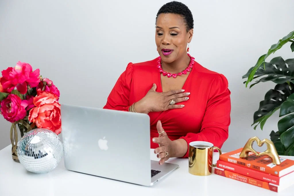 A woman in a red dress and jewelry faces a laptop, placing her hand on her chest and appearing emotional. She sits at a white desk adorned with pink flowers, books, a gold heart ornament, a coffee mug, and a disco ball, with a green plant in the back