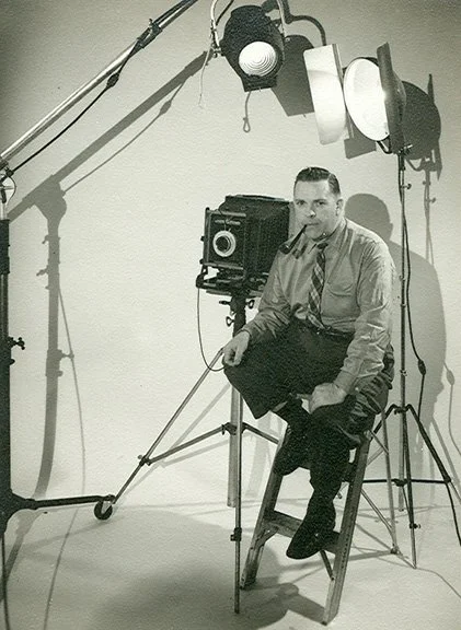 Black and white photo of a man sitting on a stool in a photography studio, surrounded by lighting and camera equipment.