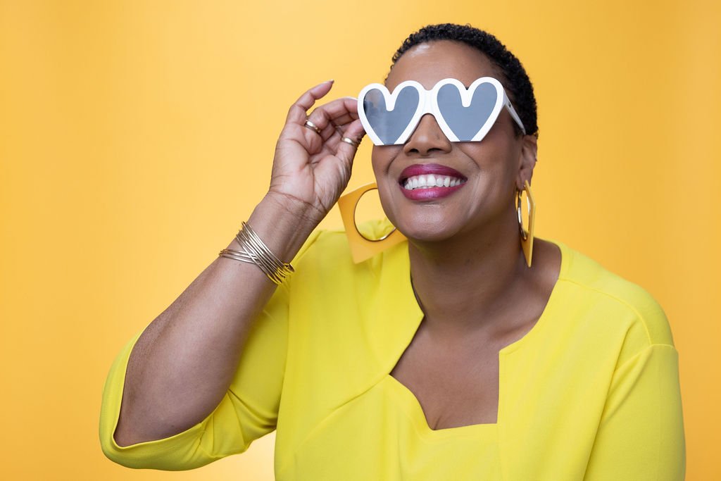 A woman wearing heart-shaped sunglasses and a yellow outfit against a yellow background, smiling and holding her glasses.