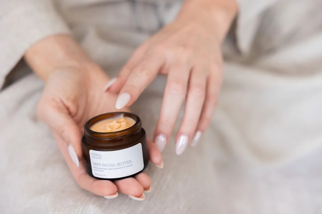 Person holding an open jar of deep facial butter cream with fingers near face, blurred background
