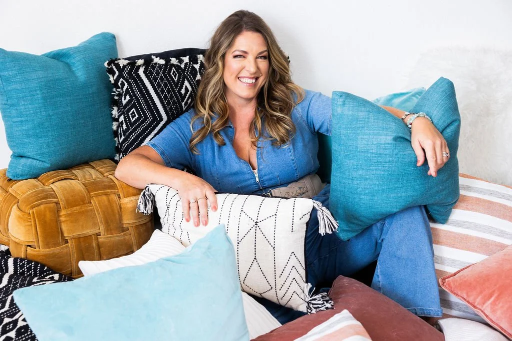 A woman sitting on a couch surrounded by colorful pillows, smiling and winking at the camera.