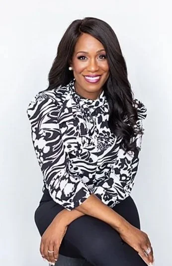 A woman with long, dark hair, wearing a black and white patterned blouse and black pants, smiling and sitting against a white background.