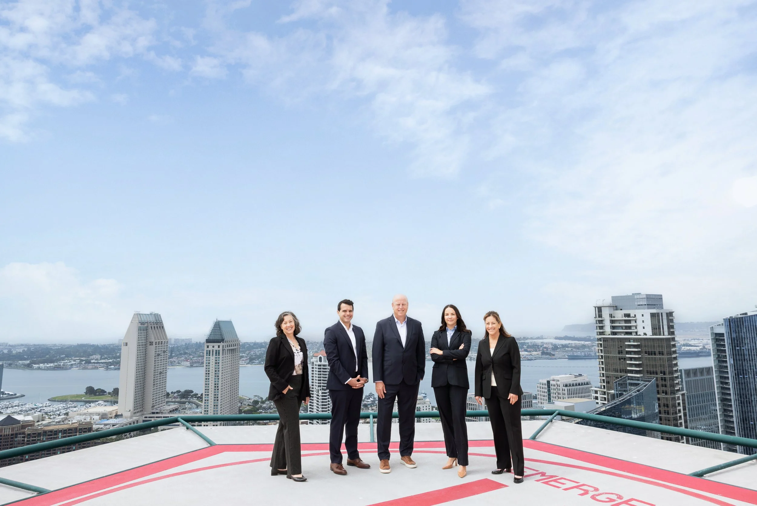 Group of five business professionals standing on rooftop helipad with city skyline and river in the background during daytime.