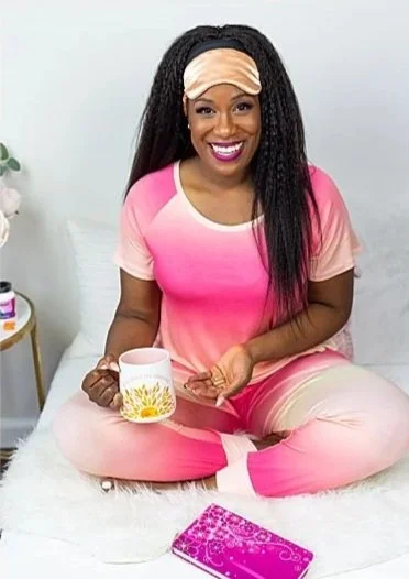 Smiling woman sitting cross-legged on bed, wearing pink pajamas and a sleep mask, holding a mug, with a pink notebook nearby