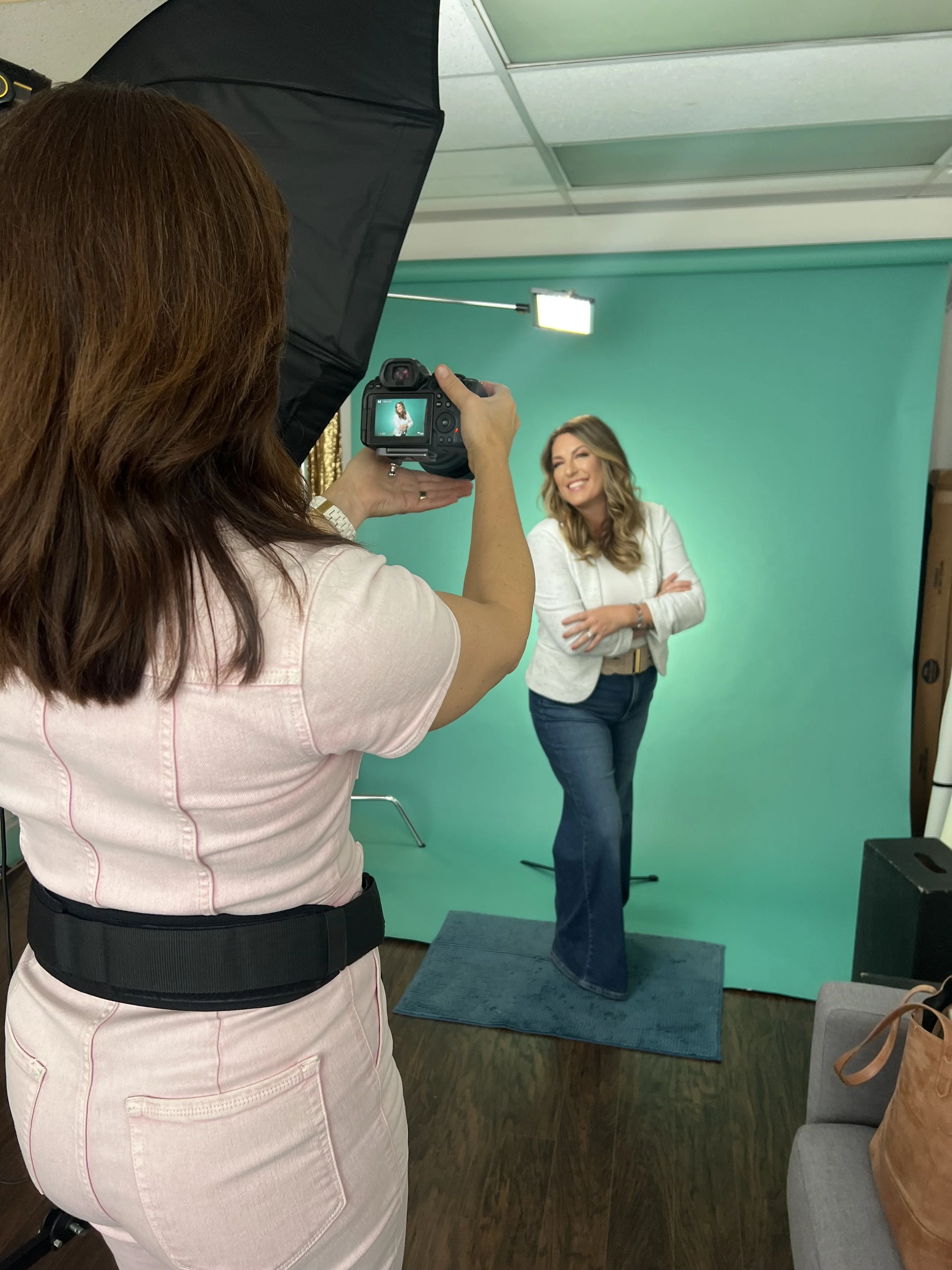 A woman with brown hair wearing a light pink shirt is photographing another woman with blonde hair smiling and standing in front of a green screen. The woman in front is dressed in a white blazer and jeans, standing on a small blue mat. There is a large softbox light overhead, and a small stool is visible in the background.