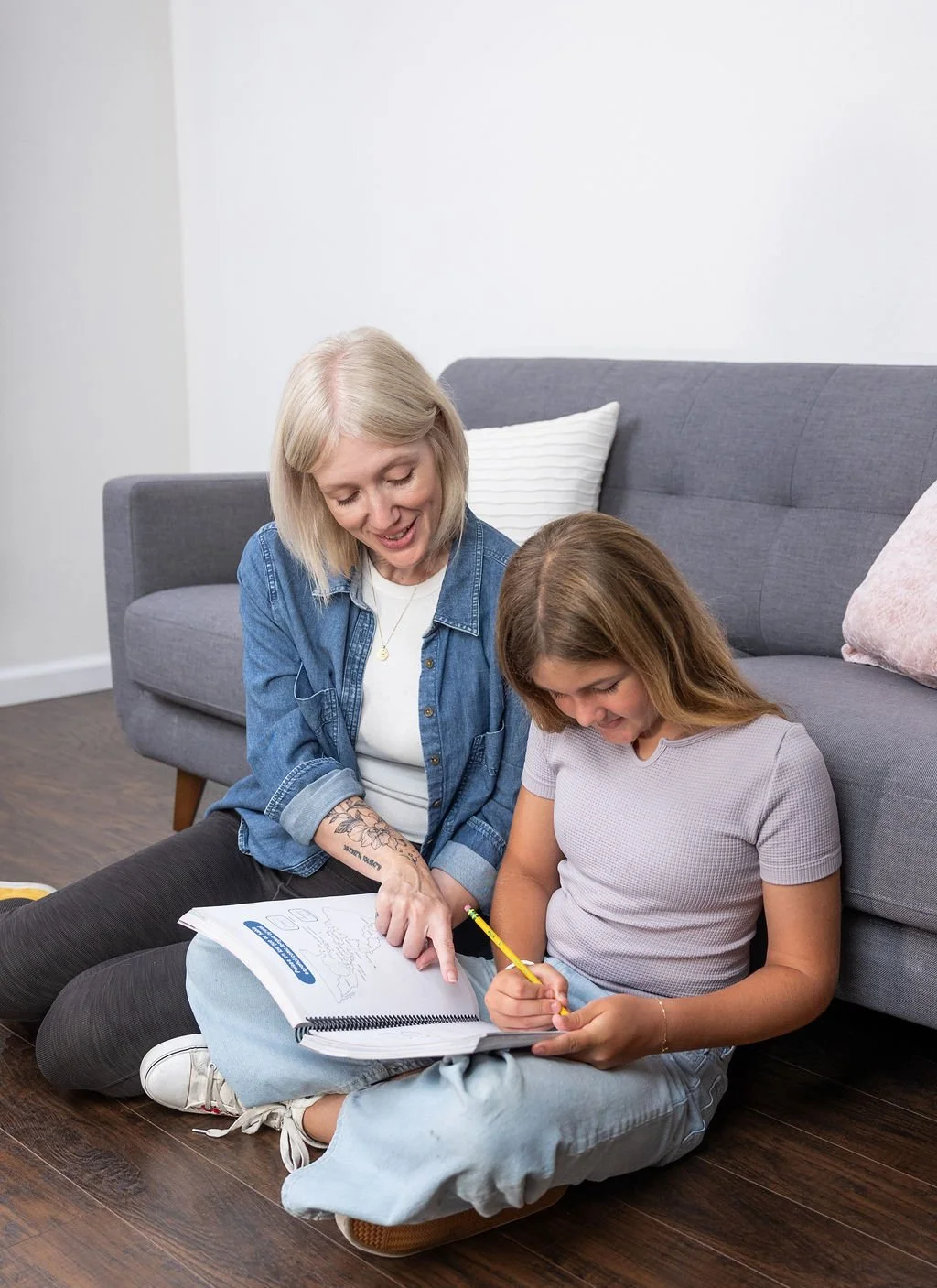 A woman and girl sitting on the floor in a living room, looking at a notebook with drawings and writing, smiling and engaged in drawing together.