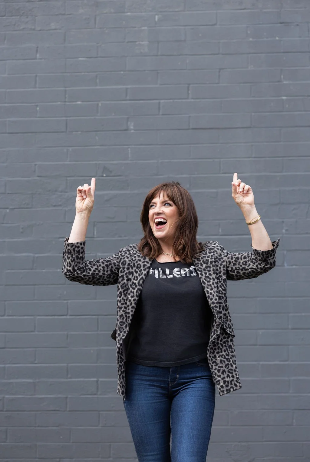 A woman smiling and laughing, standing in front of a gray brick wall, wearing a leopard print blazer and a black t-shirt with the word 'KILLER' on it, pointing upwards with both hands.