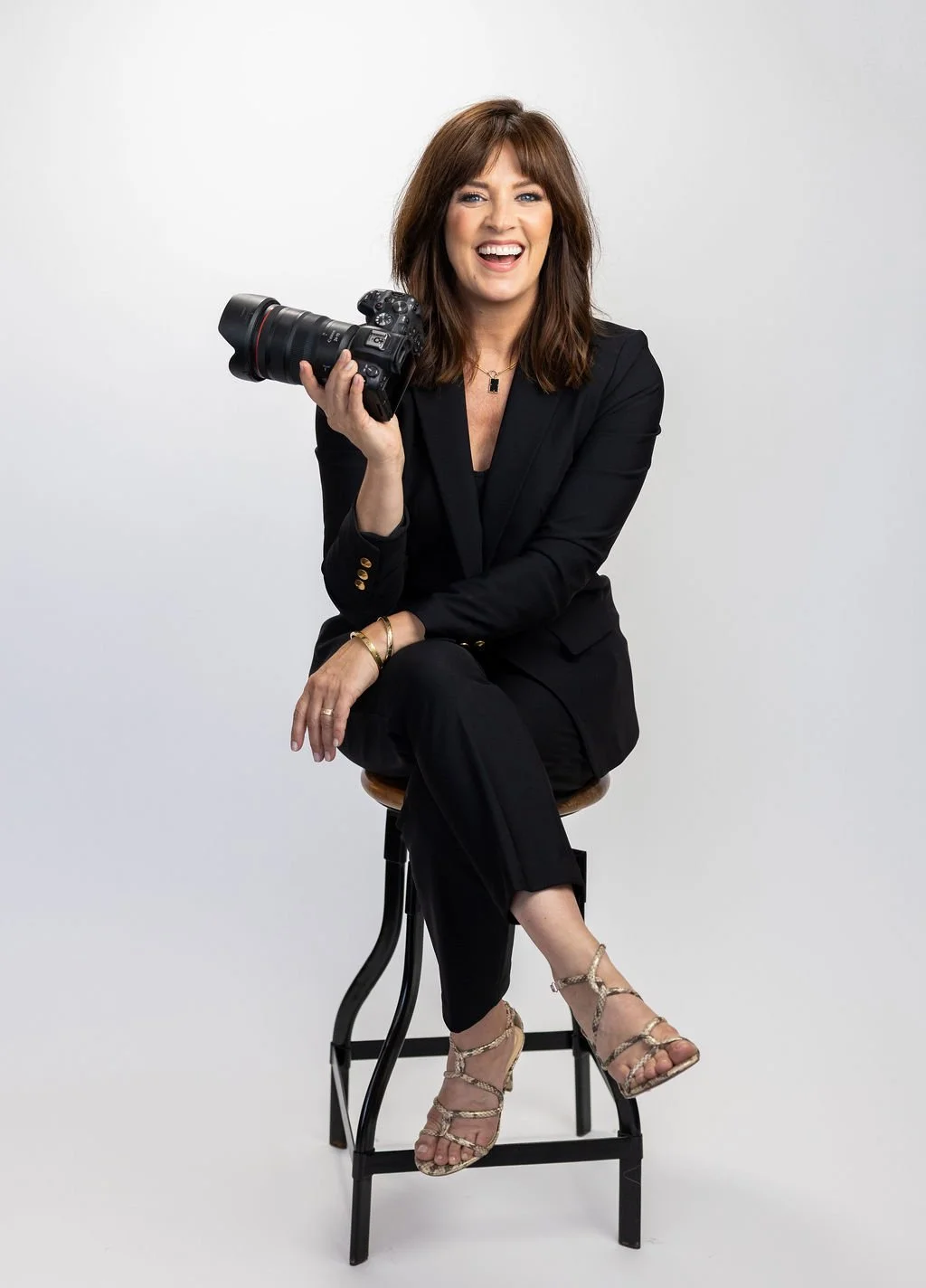 Woman in black suit holding a professional camera, sitting on a stool, smiling, on a plain white background.